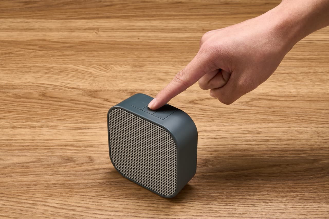 A hand pressing the top button of a slate gray Dio Node speaker on a light wood surface.