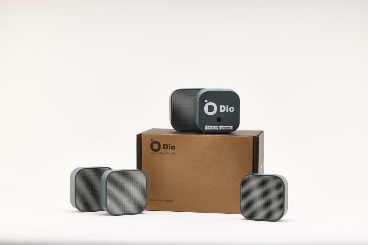 Four Dio Node speakers arranged around a kraft box printed with the Dio logo, a fifth speaker resting on top of the box.