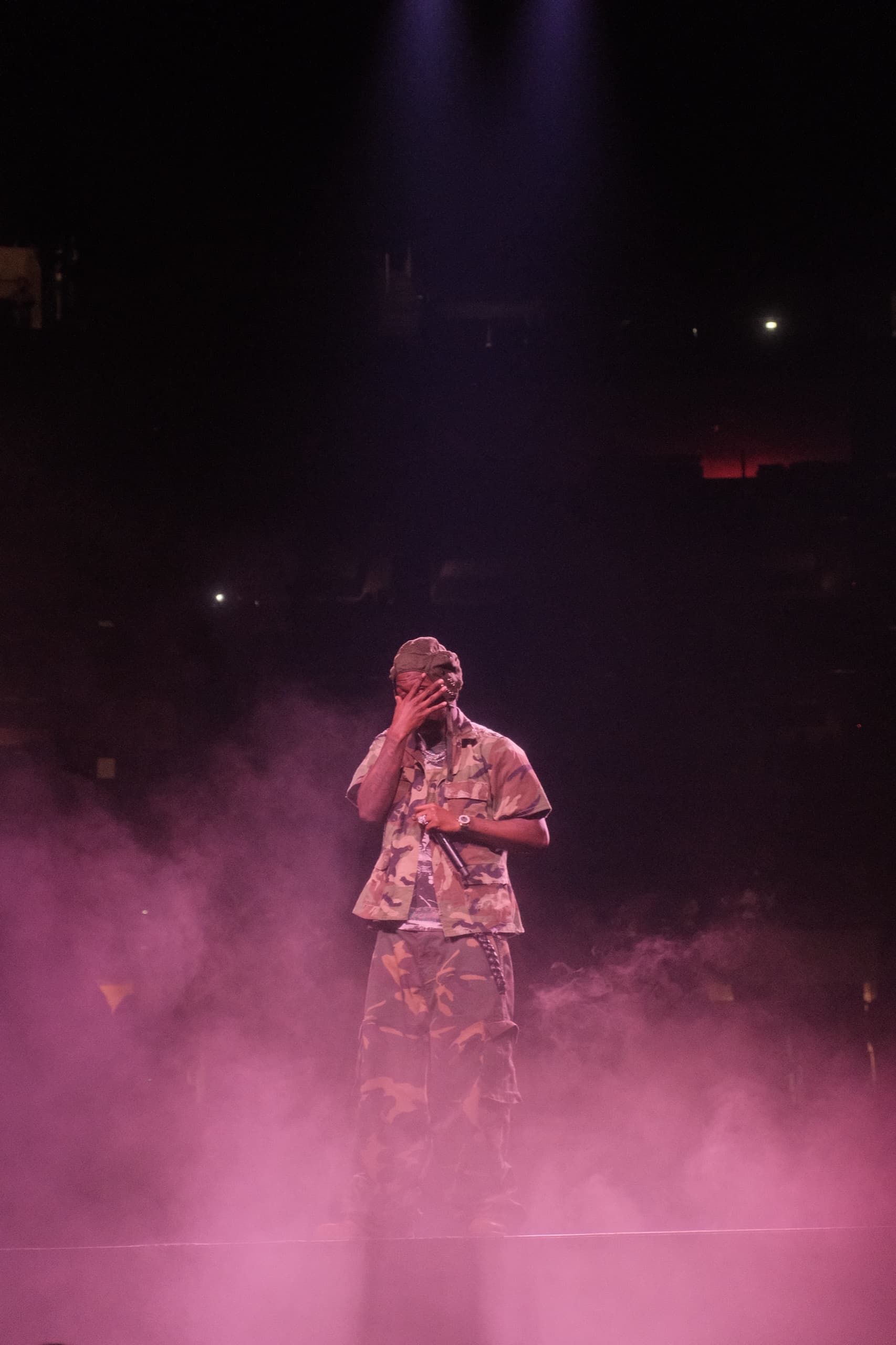 A performer in a patterned shirt sings into a handheld mic, lit by magenta haze against a dark stage.