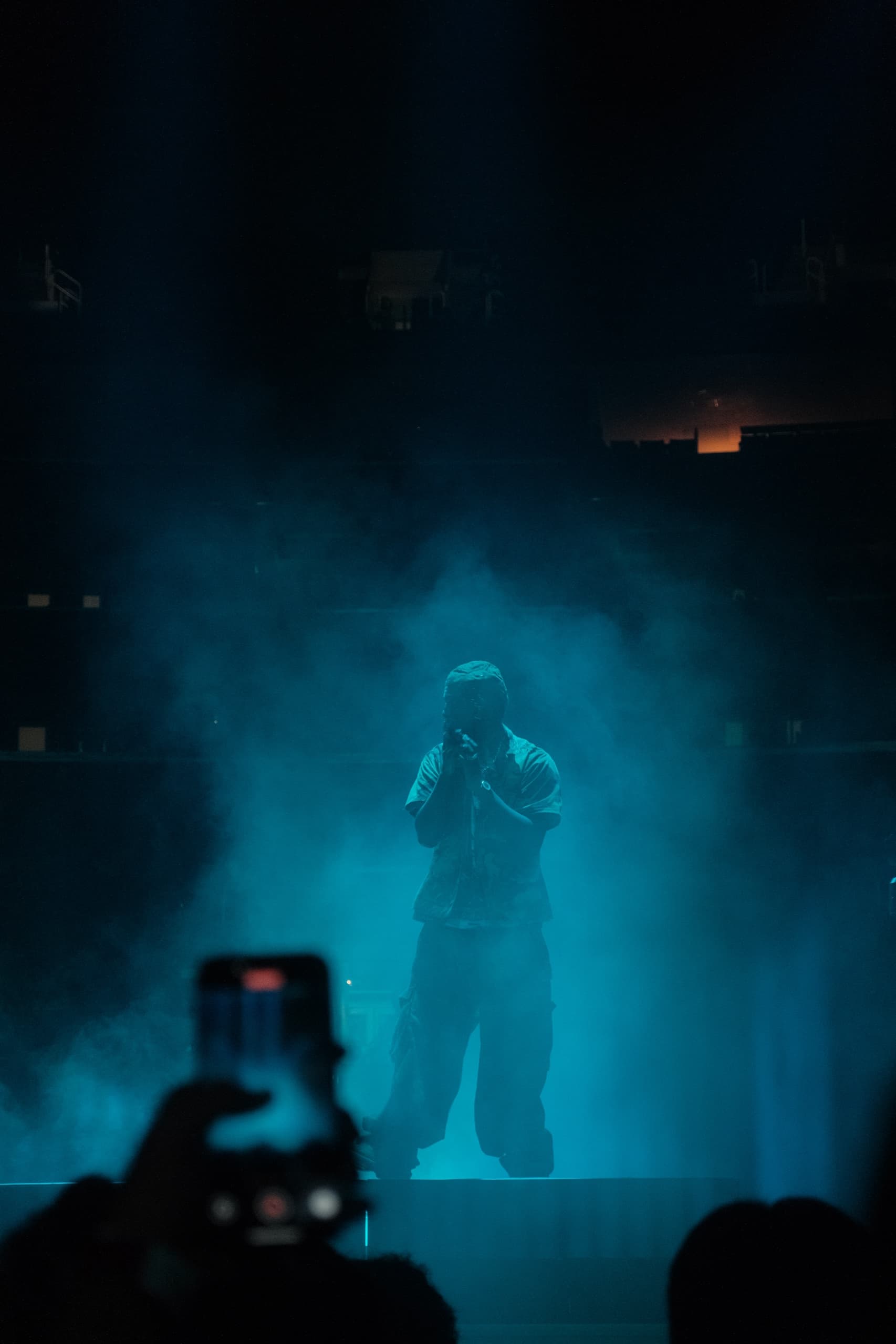 A performer silhouetted at the mic in heavy teal stage haze, with raised hands and a glowing phone screen in the crowd below.