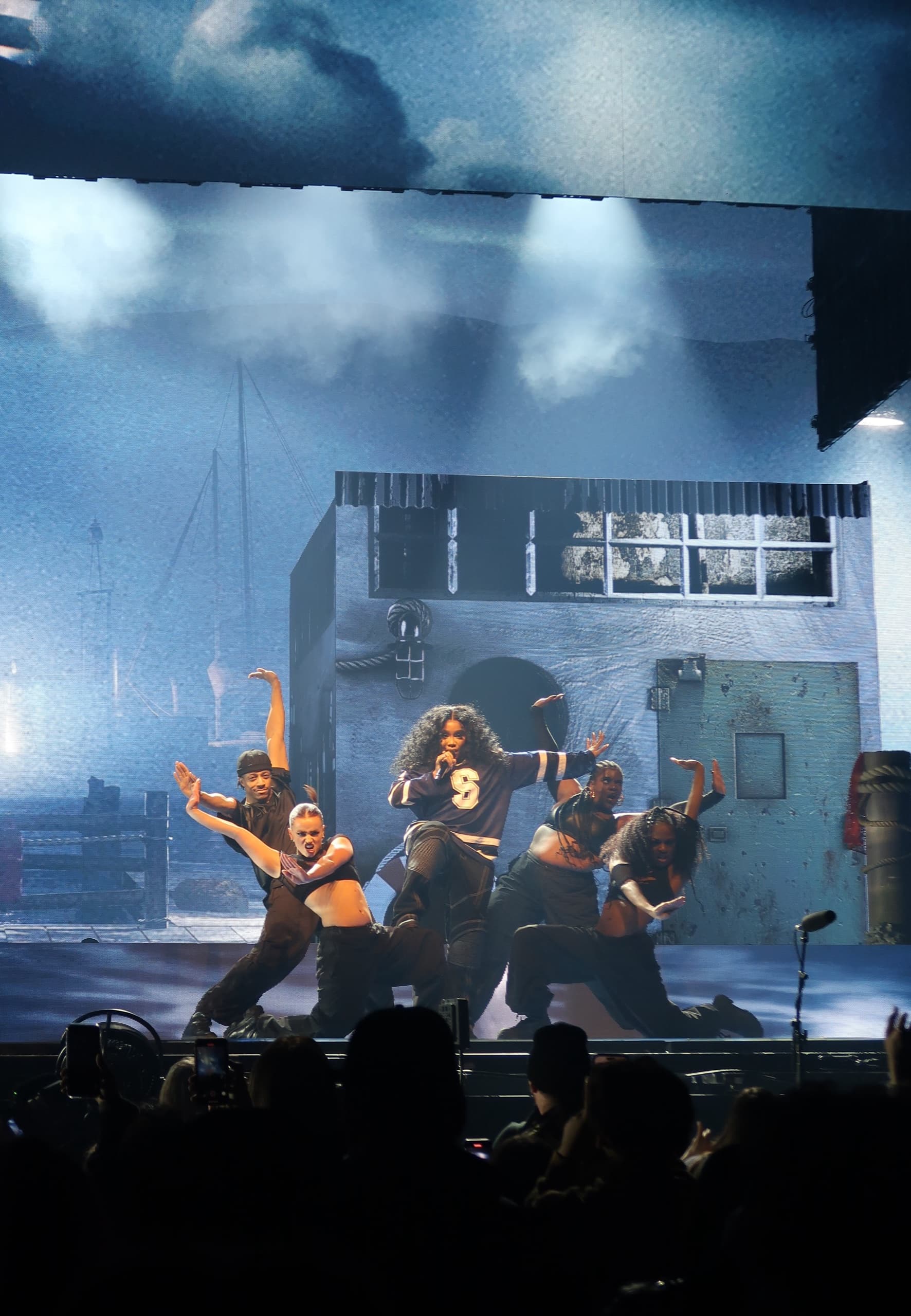Dancers performing in front of a boathouse stage set under blue light, the crowd silhouetted along the front of the stage.