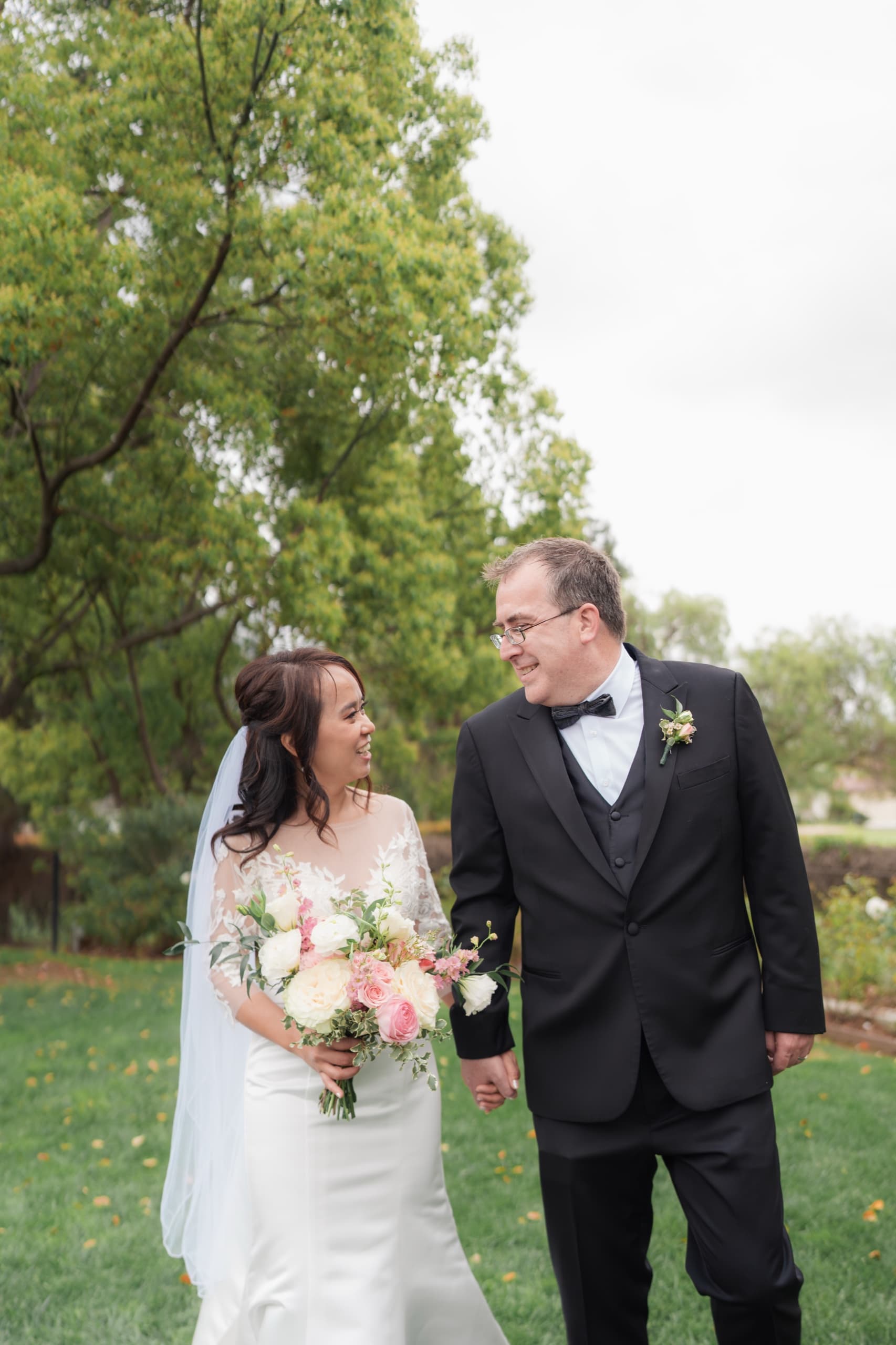 A bride and groom walk hand in hand across a lawn, looking at each other, her bouquet of white and blush roses at her side.