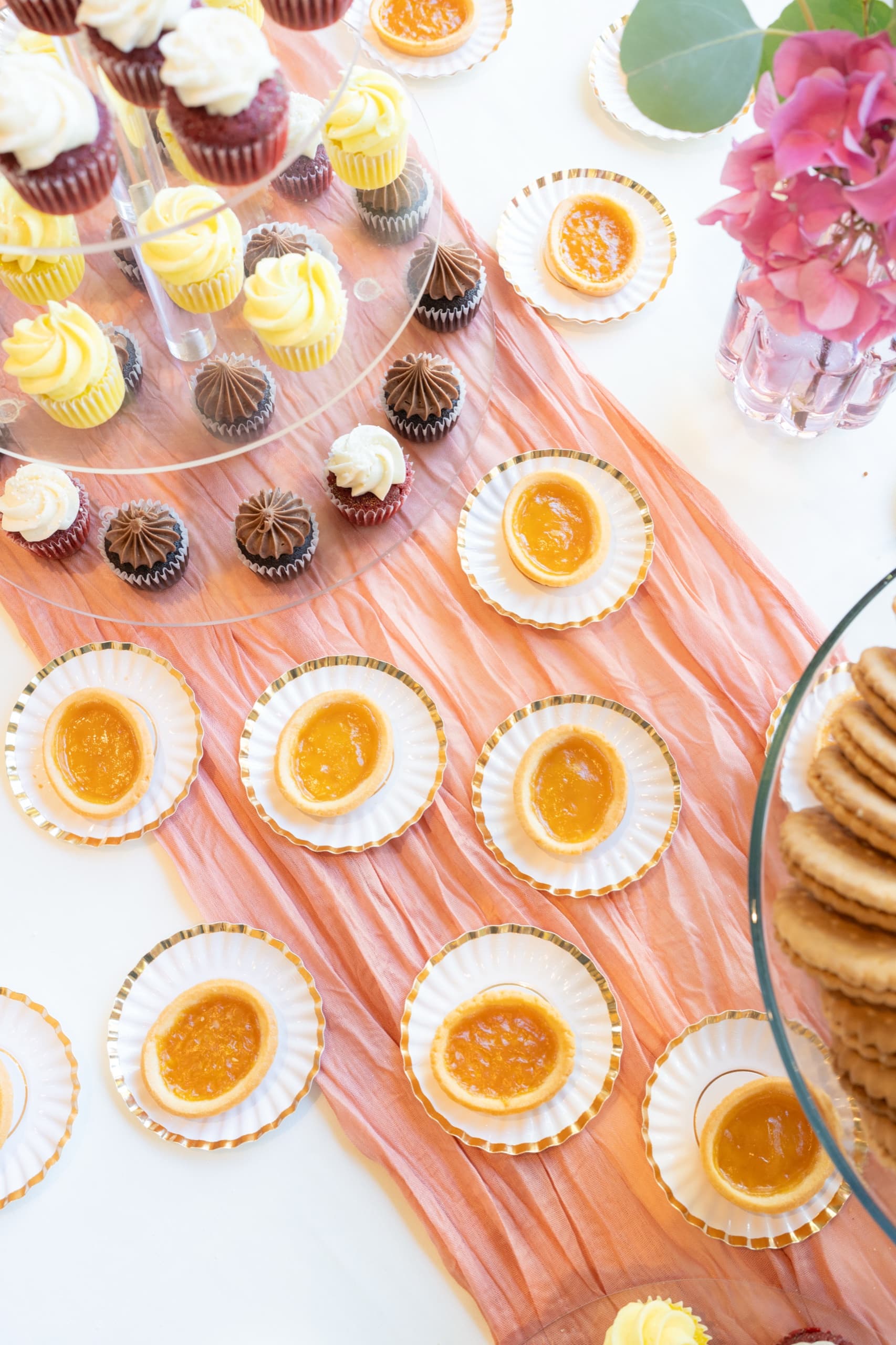Cupcakes on a glass stand beside rows of lemon tarts on gold-rimmed plates, arranged on a peach gauze runner.