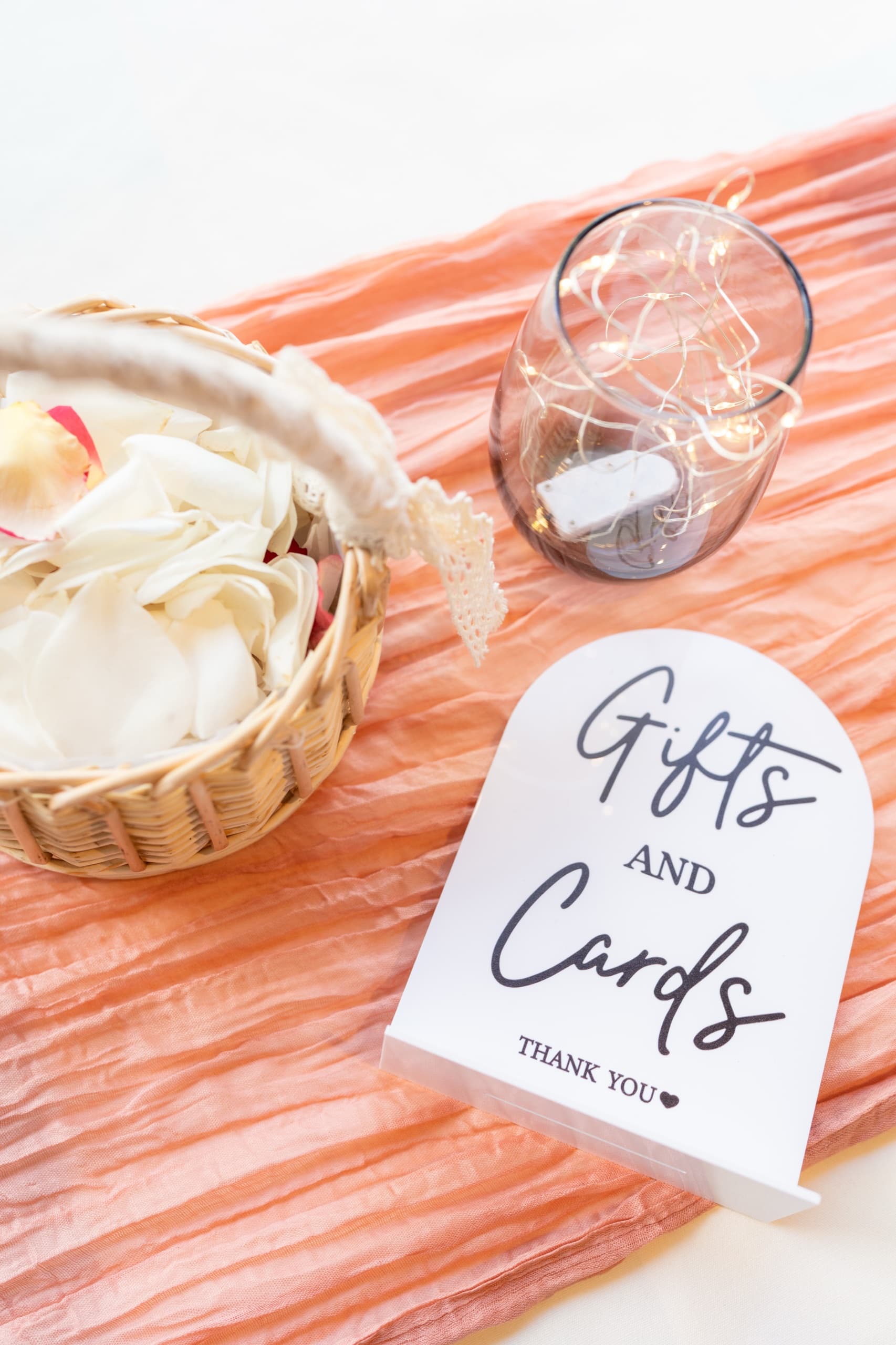 A gifts and cards sign beside a basket of ivory petals and a glass of fairy lights on a peach runner.
