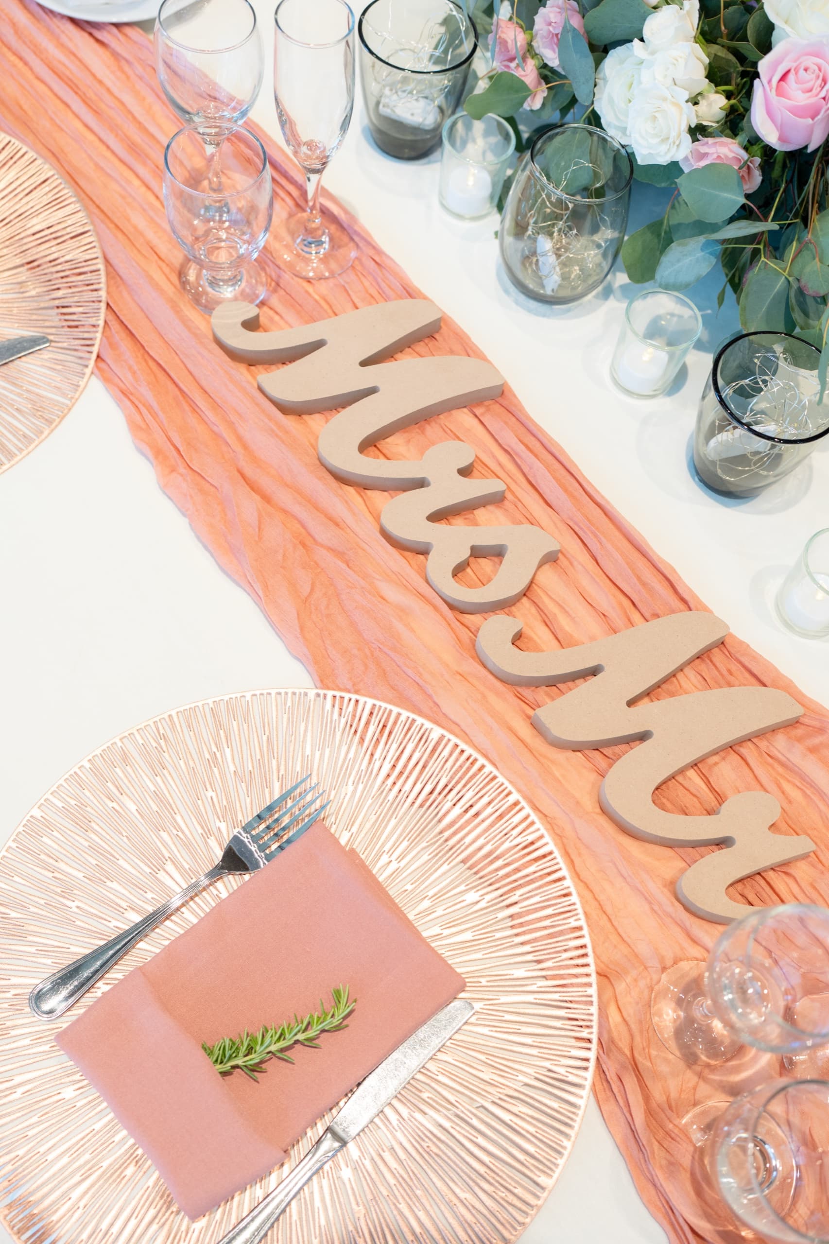 A reception table set with rose-gold charger plates, blush napkins topped with rosemary, and wooden Mr and Mrs letters along a peach runner.