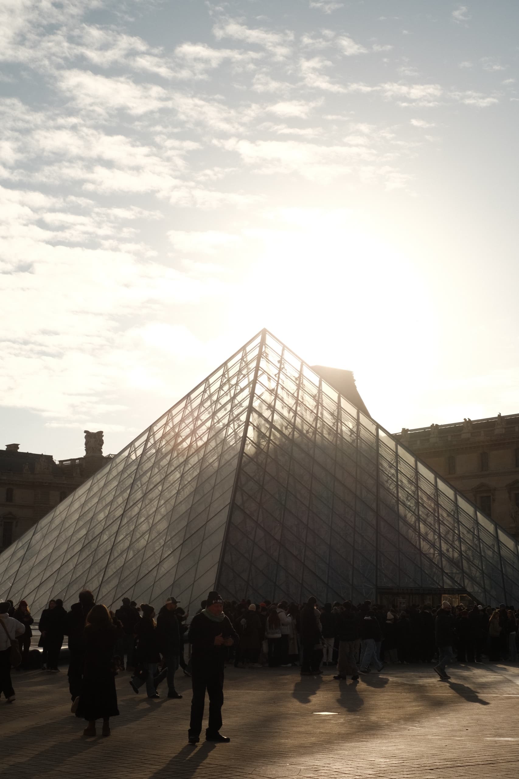 Crowds crossing the Louvre courtyard at low sun, the glass pyramid backlit against a bright sky.