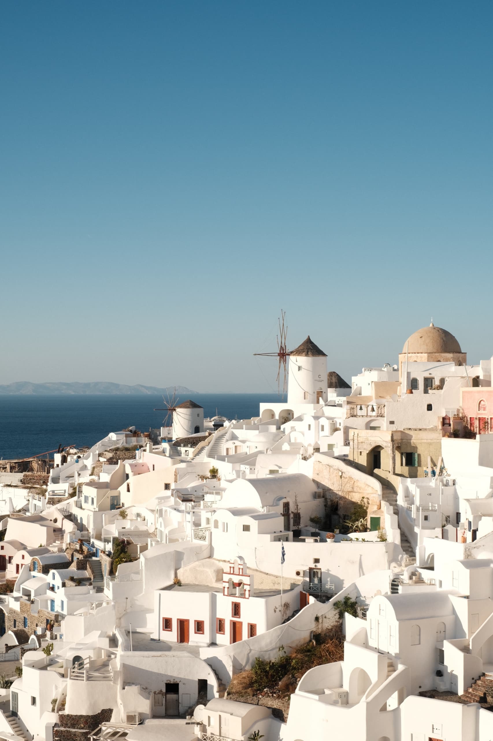 A whitewashed Greek island village stepping down a hillside to the sea, windmills and a domed church in warm light.