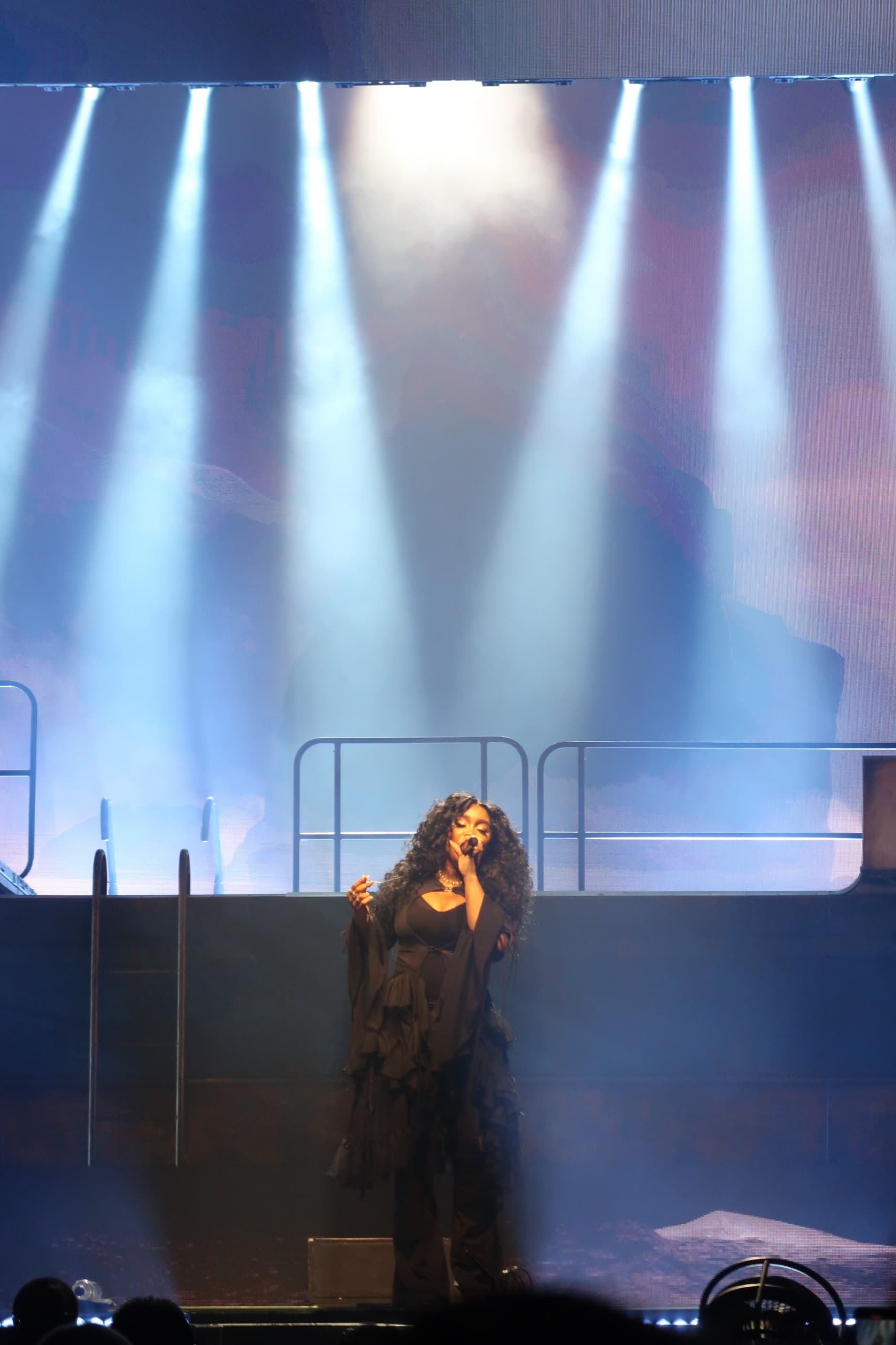 A singer alone at center stage under a row of blue spotlight beams cutting down through haze.