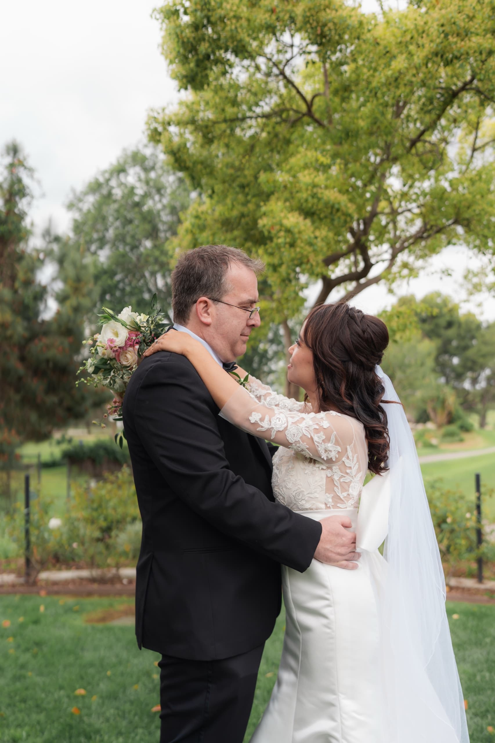 A bride with her arms around the groom's shoulders on a garden lawn, her bouquet raised behind him and her veil trailing.